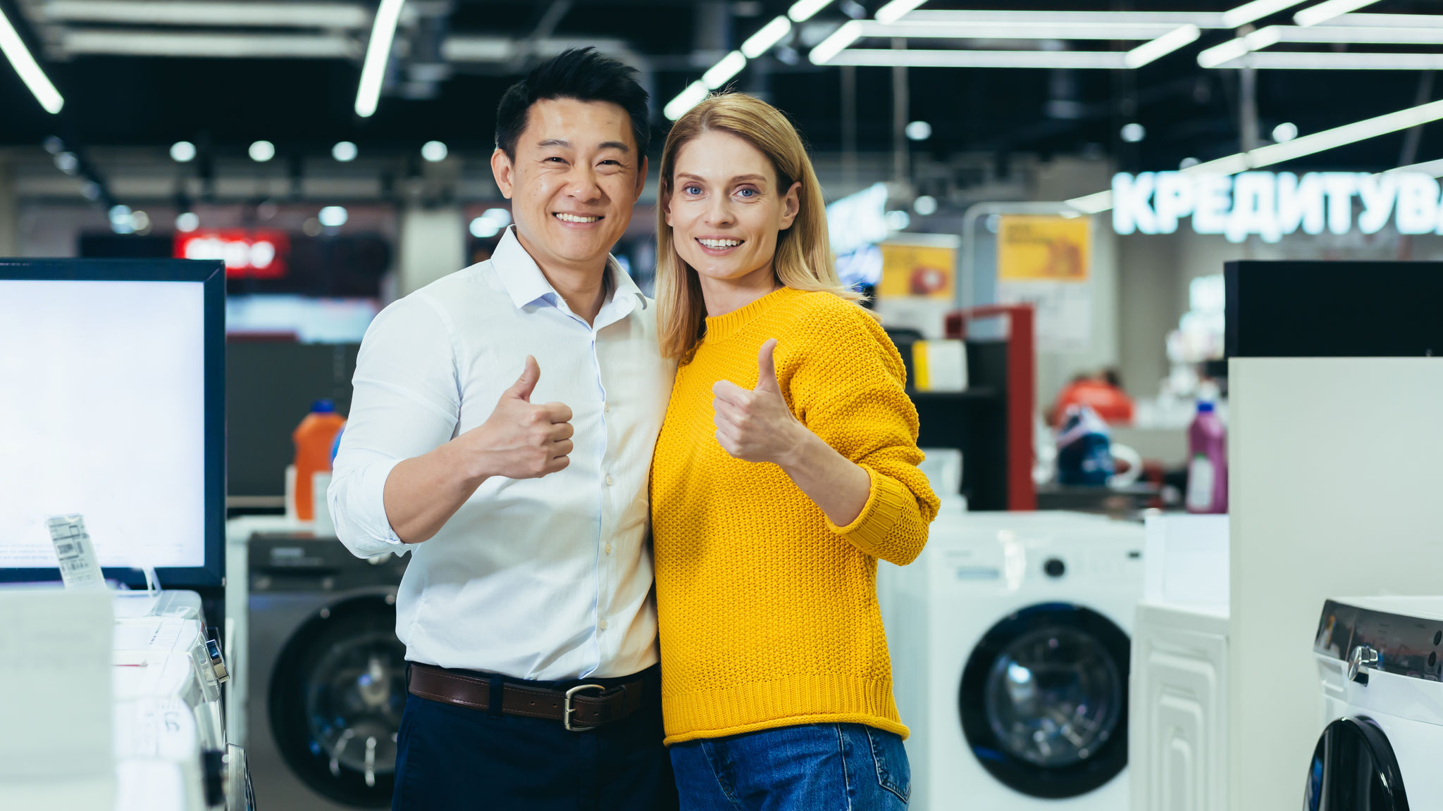 Nakangiting mag asawa na may hawak na thumbs up sa harap ng isang electronic store 