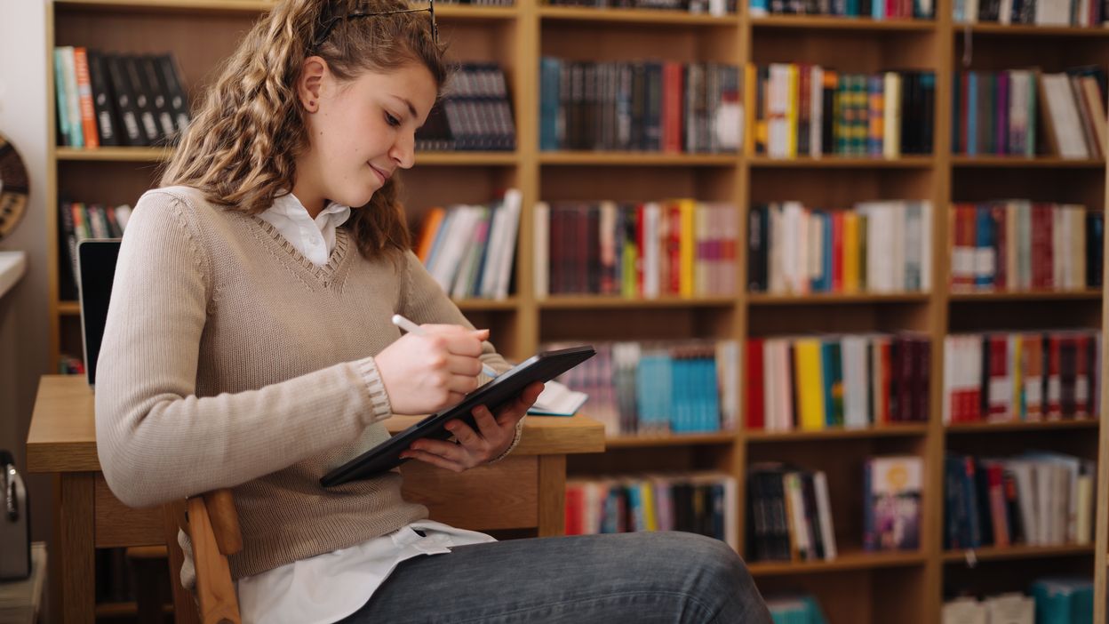 woman-drawing-on-an-ipad-while-sitting-in-a-bookst-2022-11-16-11-41-40-utc