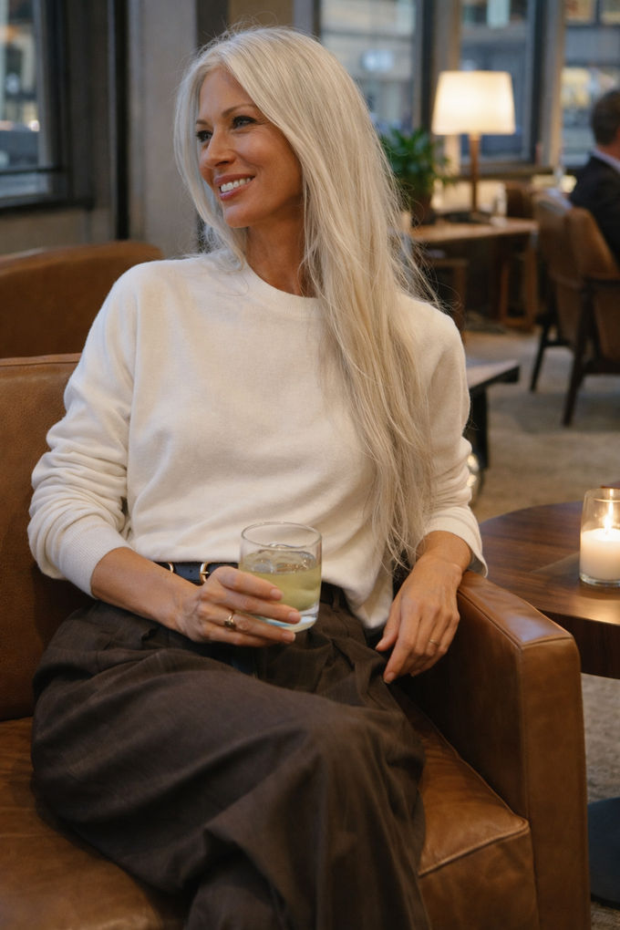 Woman seated in a modern lounge wearing a cream knit sweater and dark high-waisted trousers, smiling while holding a drink, cozy office fashion.