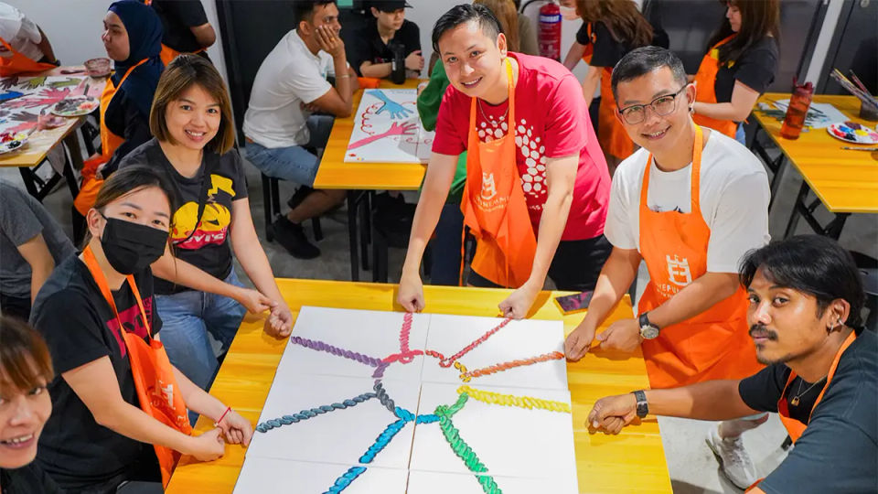 Painters around a long table working on a bright collaborative canvas