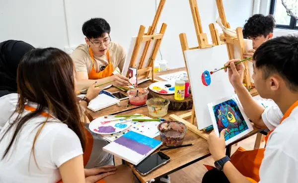 Young colleagues in orange aprons paint canvases on easels around a table, a phone propped up beside them
