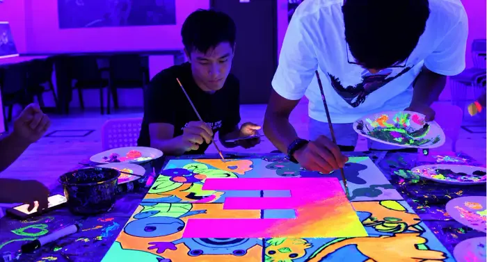 Two participants paint a large neon canvas under UV light