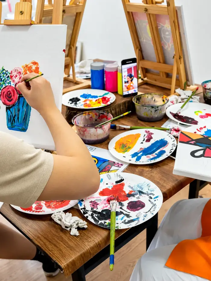A participant paints flowers on a canvas, palettes of acrylic paint beside it
