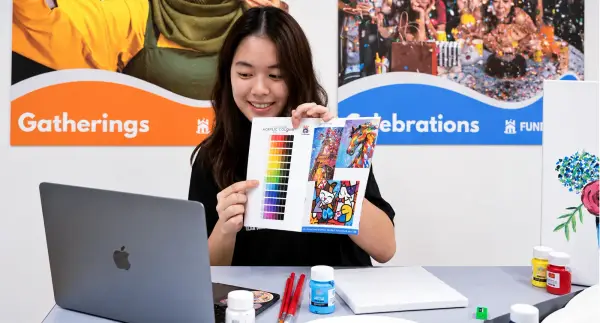 A facilitator leads a virtual art jamming session from a laptop, holding up a colour chart