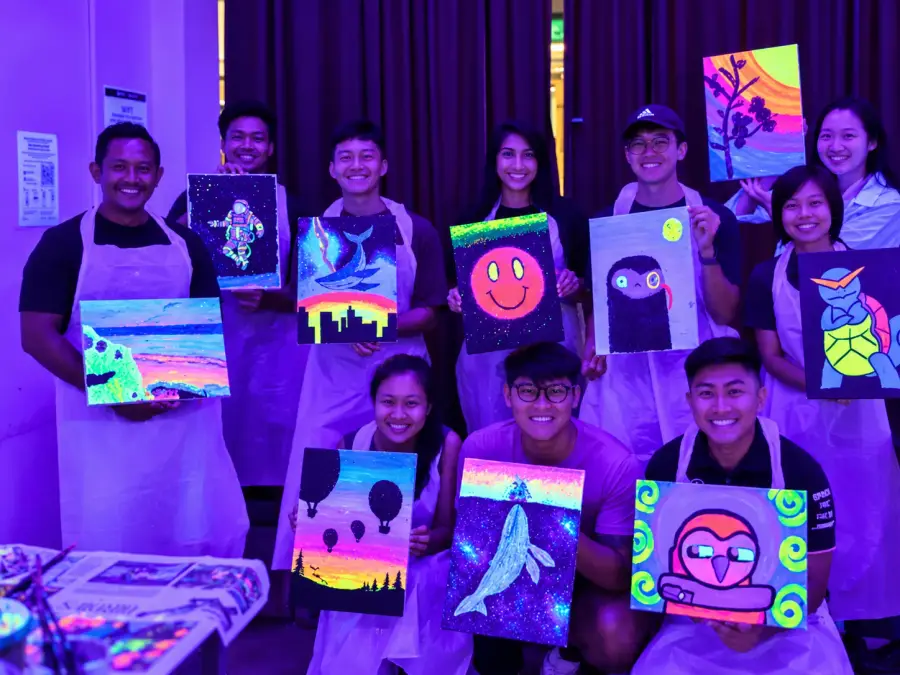 A group hold up the neon paintings they each made, glowing under UV light