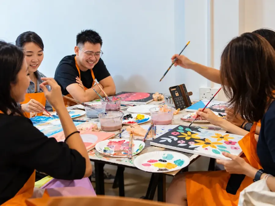 A team paint their canvases around a table, with a phone showing a locked puzzle propped up between them