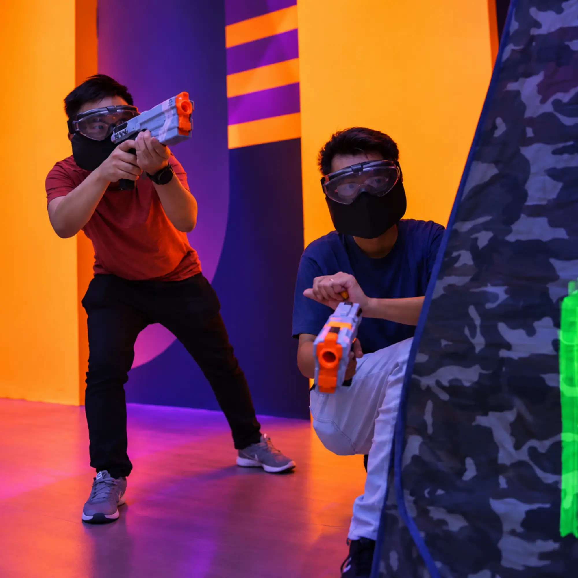 Two players in masks take aim with foam blasters beside a bunker in the studio