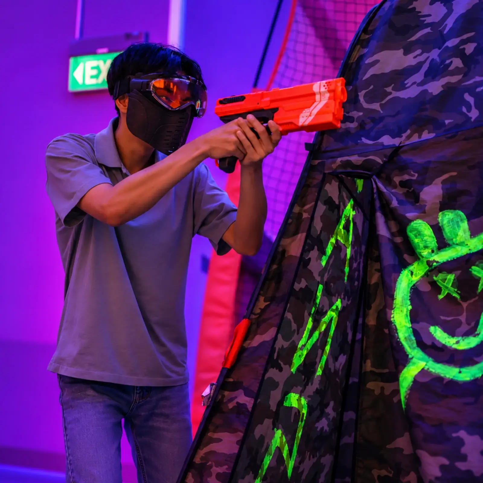 A player in a mask and goggles aims a foam blaster from behind a bunker