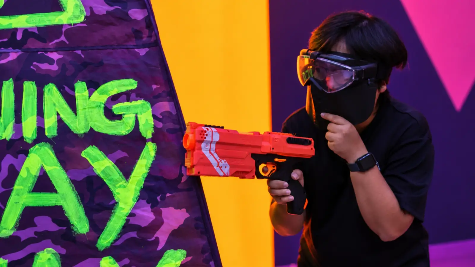 A player in a mask aims a foam blaster from behind a neon-painted bunker