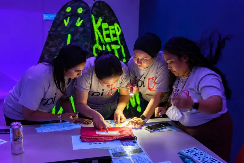 Four players crowd over a clue under a glowing neon sign