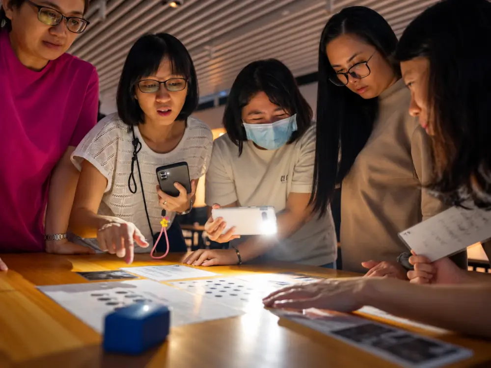 Five players lean over a lit puzzle sheet, one pointing at a clue