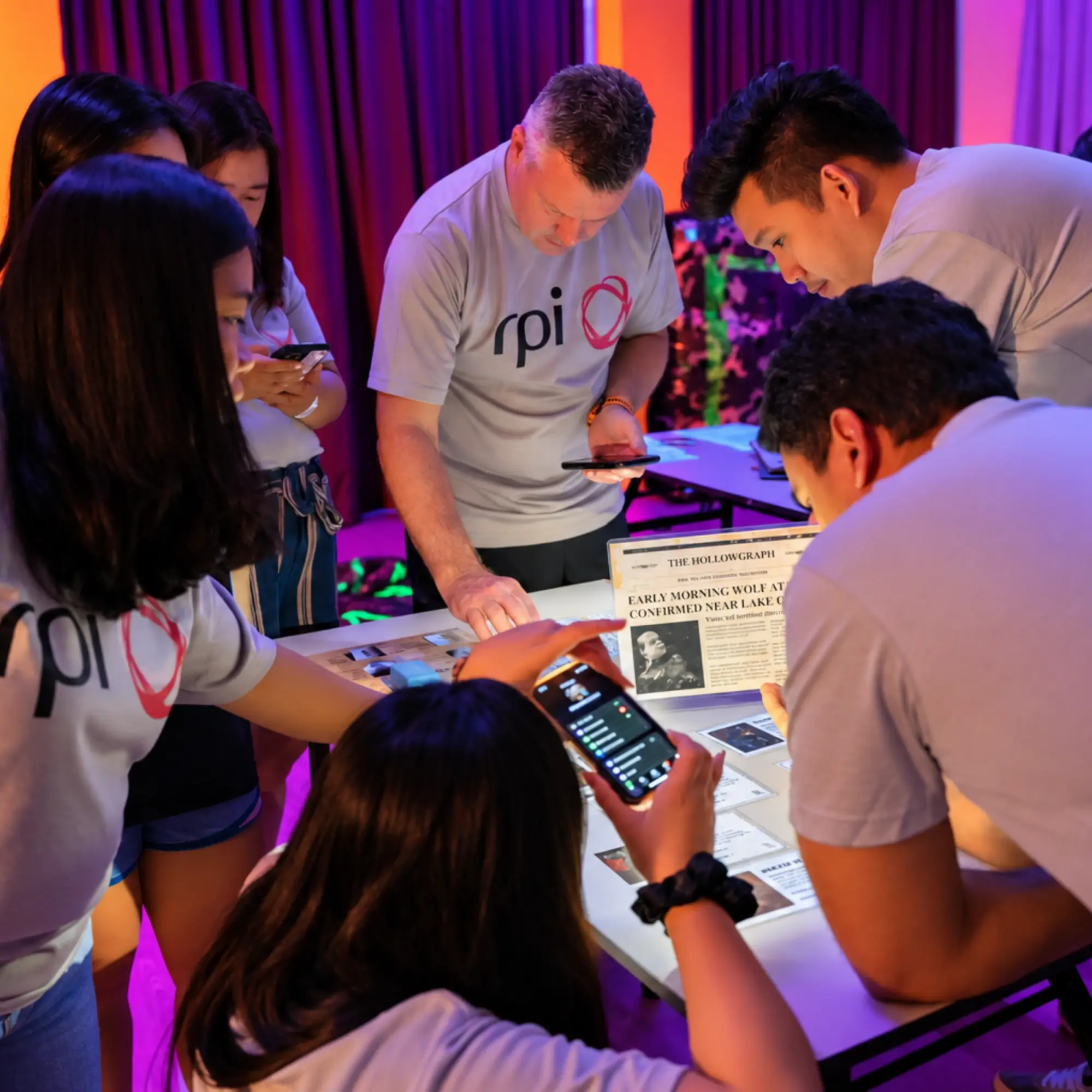 A team in matching shirts leans over a table of clues, one holding a phone over a newspaper prop