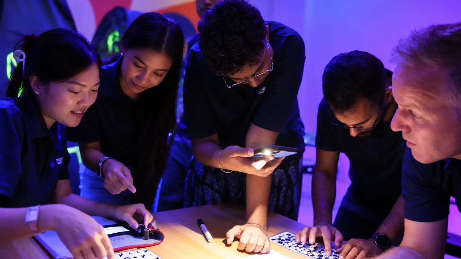Five teammates lean in over a puzzle in a darkened room, their faces lit by a phone torch