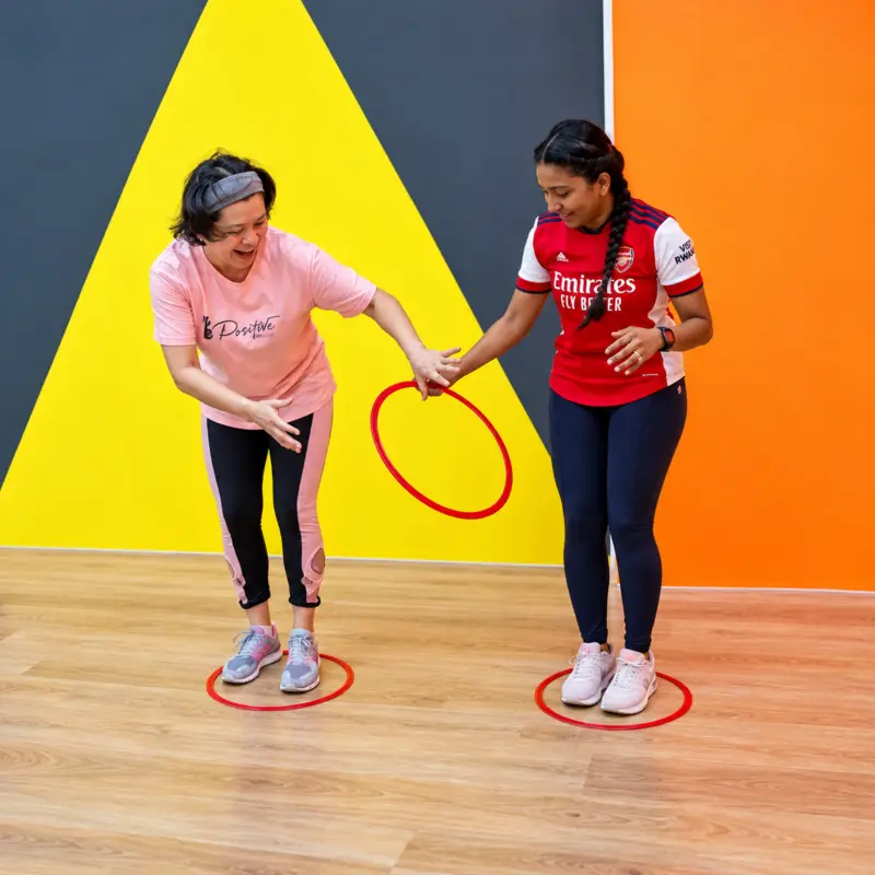 Two players, each standing in a floor ring, pass a hula hoop over their joined hands