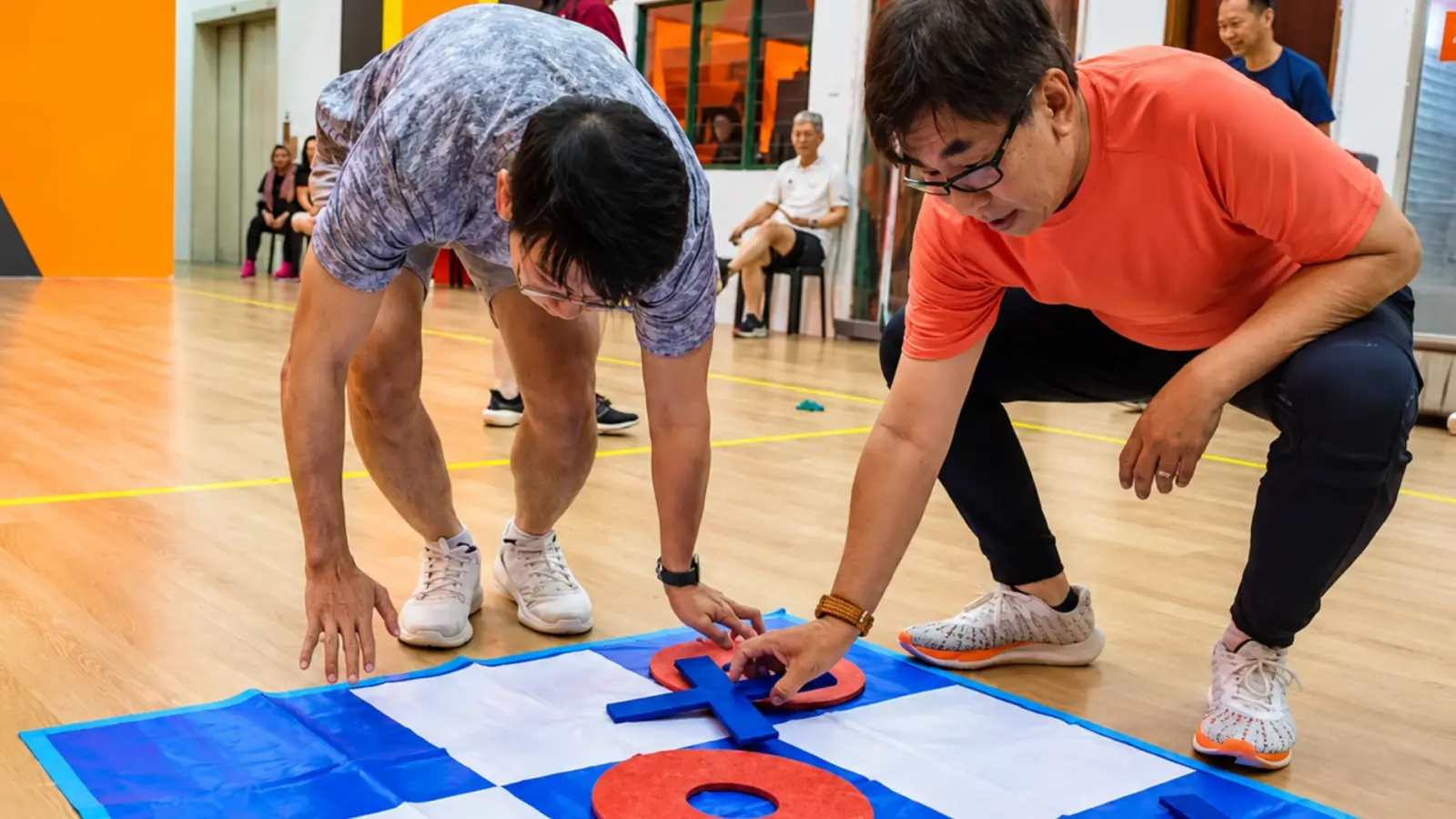 Two players crouch over a giant noughts-and-crosses mat, placing a cross
