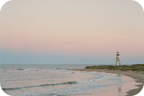 Pastel sunset over a quiet sandy beach with gentle waves, foreground cacti, and a lone lighthouse on a distant dune.
