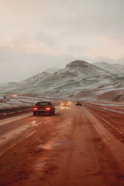 Cars drive along a wet, muddy road through snow-covered mountains at dusk.