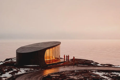 Curved modern pavilion with warm glass facade beside a winding wet road on a snowy coastal cliff at dusk as four people look out.
