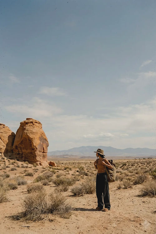 Hiker with a backpack stands in a desert landscape beside red rock formations with distant mountains under a blue sky.