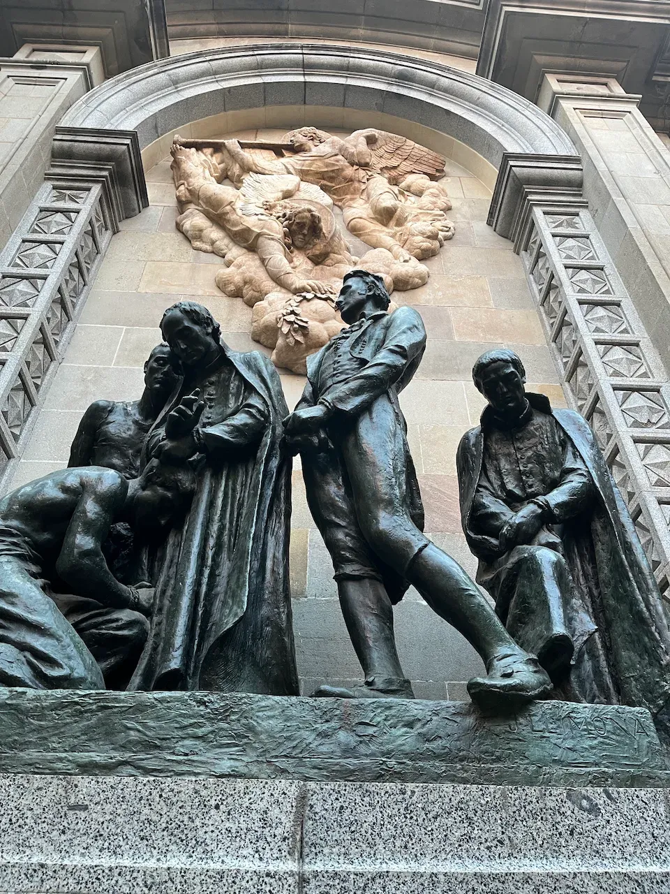 Bronze sculpture of five kneeling and standing figures beneath a stone angel relief in a Barcelona archway.