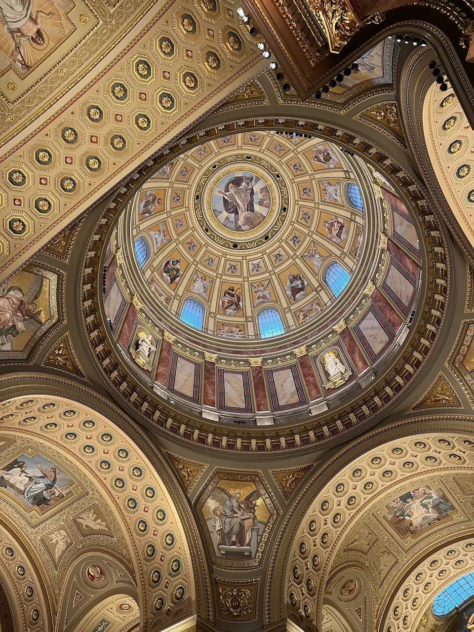 Low-angle view of a gilded cathedral dome with frescoes, statues, and arched windows.