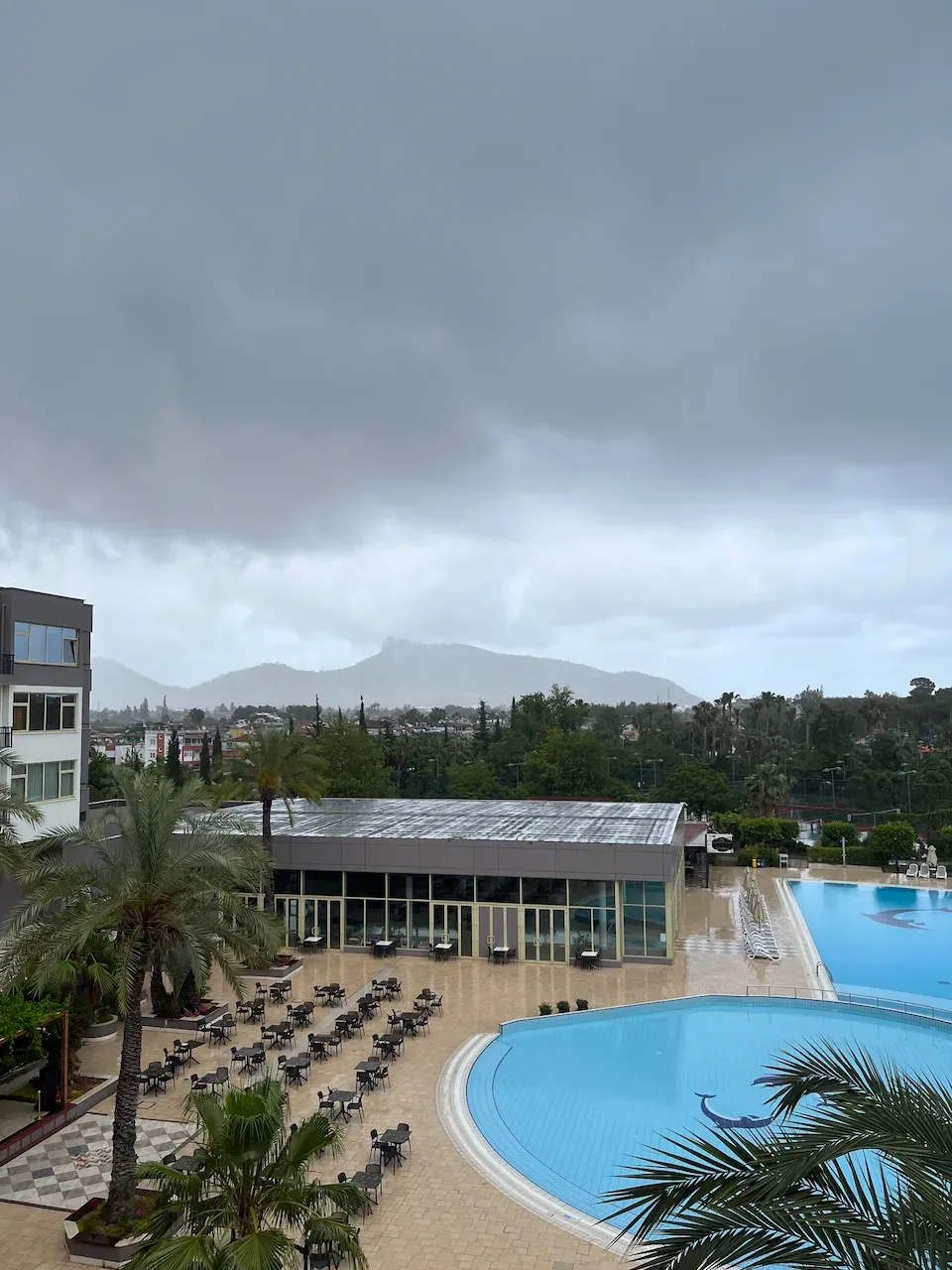 Resort swimming pools and empty patio seating under a dark overcast sky with misty mountains.