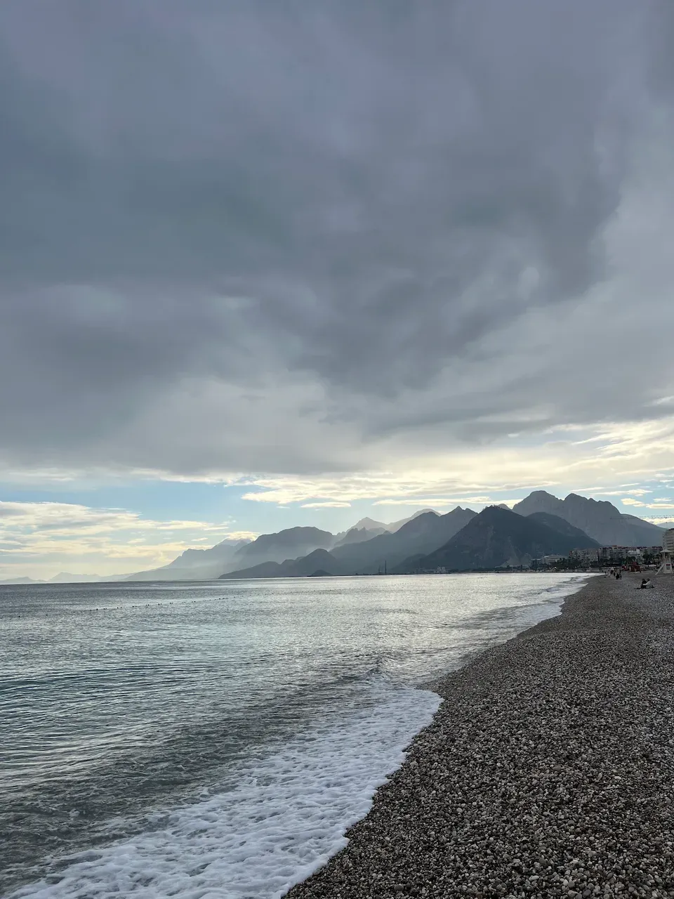 Pebble beach at dusk with calm waves, distant mountains, and a dramatic cloudy sky.