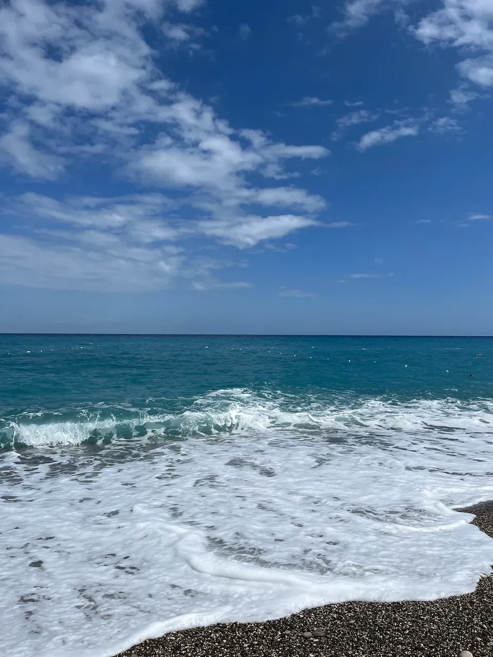 Turquoise sea waves with white foam washing onto a dark pebble beach under a blue sky.