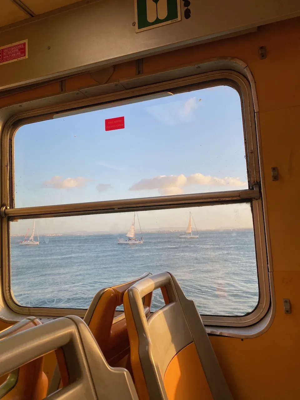View from a ferry window of three sailboats on calm blue water at golden hour.
