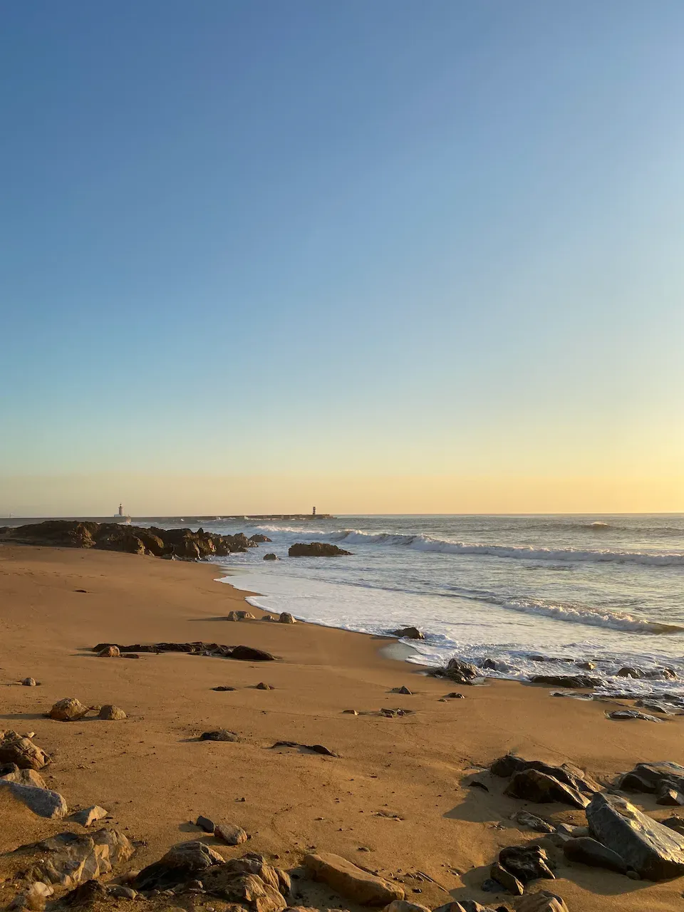 Wide sandy beach with scattered rocks and gentle waves at golden hour, with a distant lighthouse.
