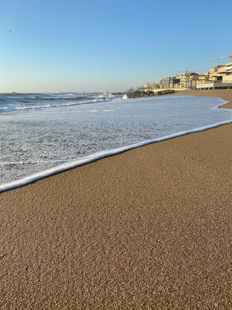Sandy beach with sea foam and waves, with a coastal city and construction cranes in the background.