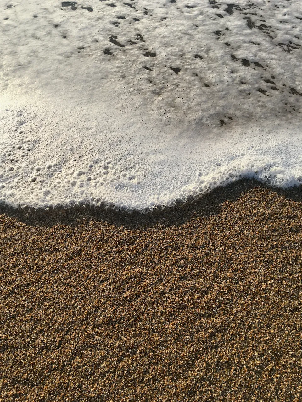 Close-up of white bubbly sea foam washing over golden-brown sandy beach.