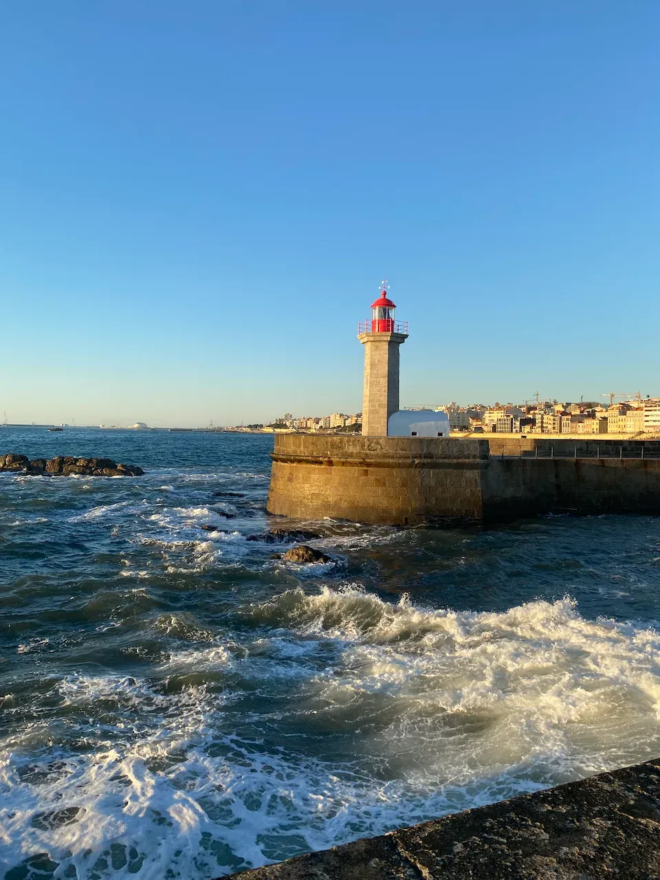 Stone lighthouse on a breakwater with crashing waves and a coastal city behind.