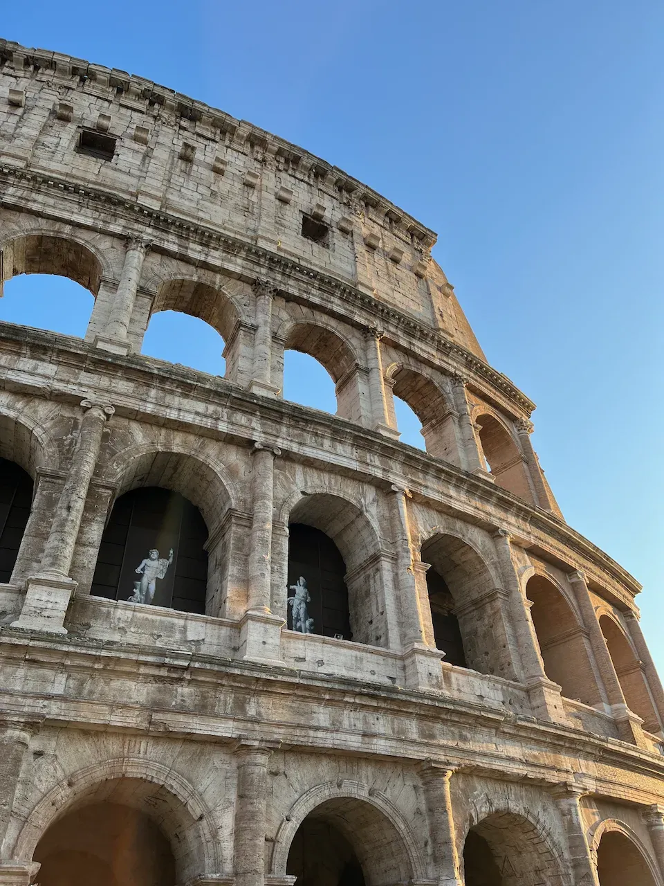 Low-angle view of the Colosseum's weathered stone arches and statues under a clear blue sky.