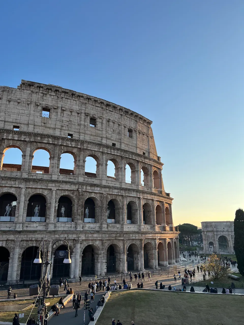 Wide view of the Colosseum at golden hour with the Arch of Constantine and tourists below.