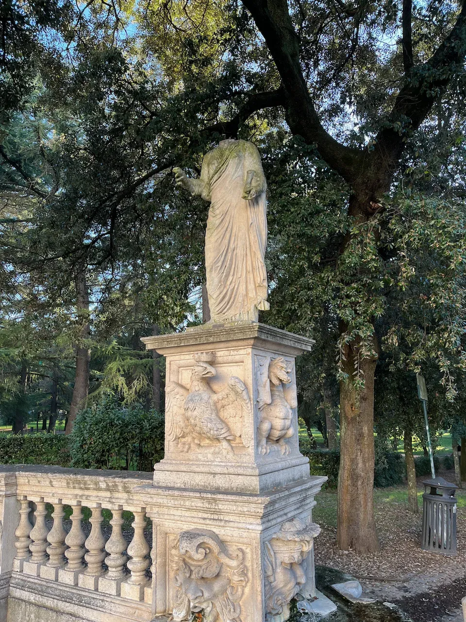 Headless classical stone statue on an ornate pedestal in a tree-lined park.