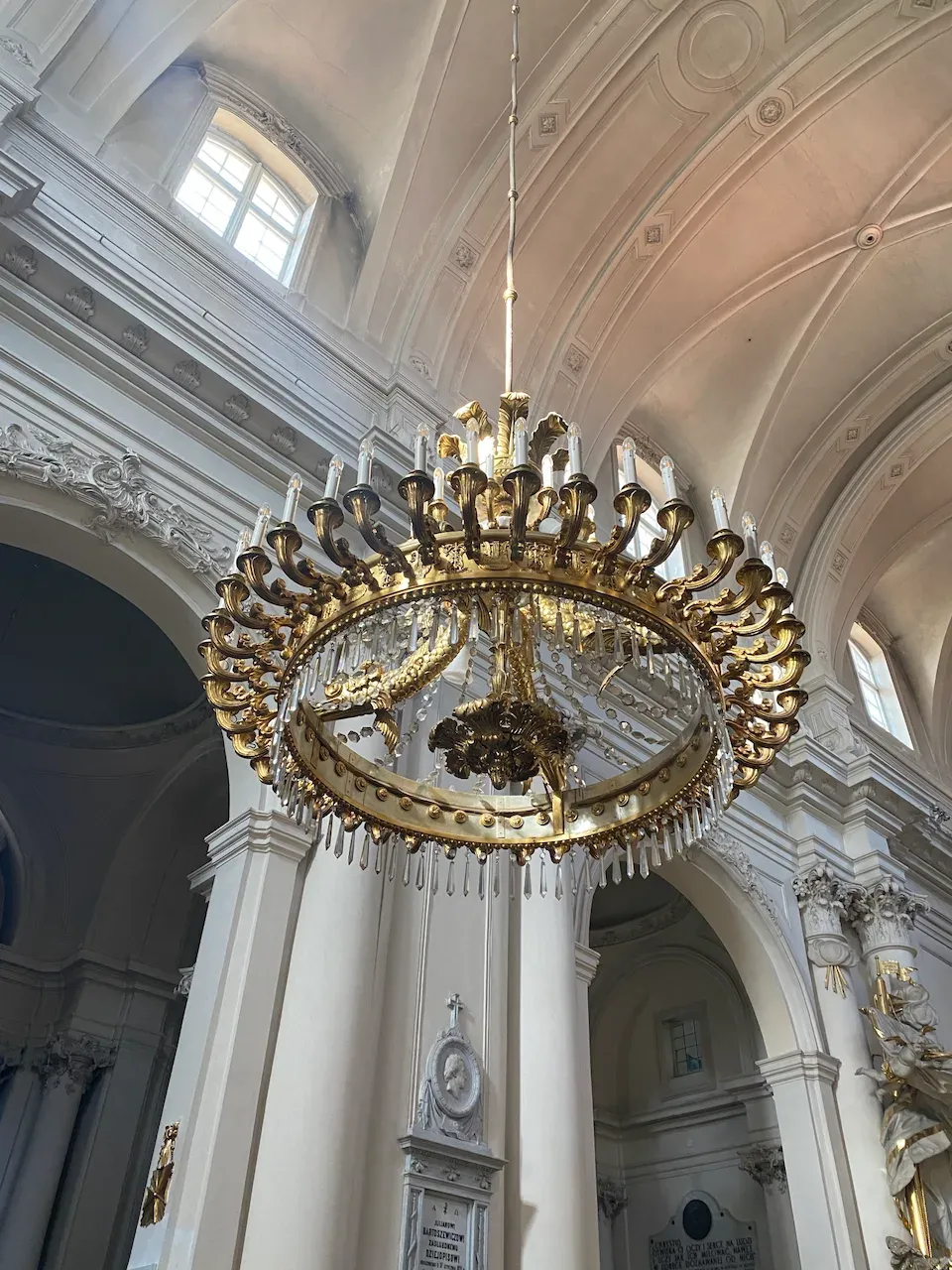 Large gold chandelier with crystal pendants hanging in a white Baroque cathedral interior.