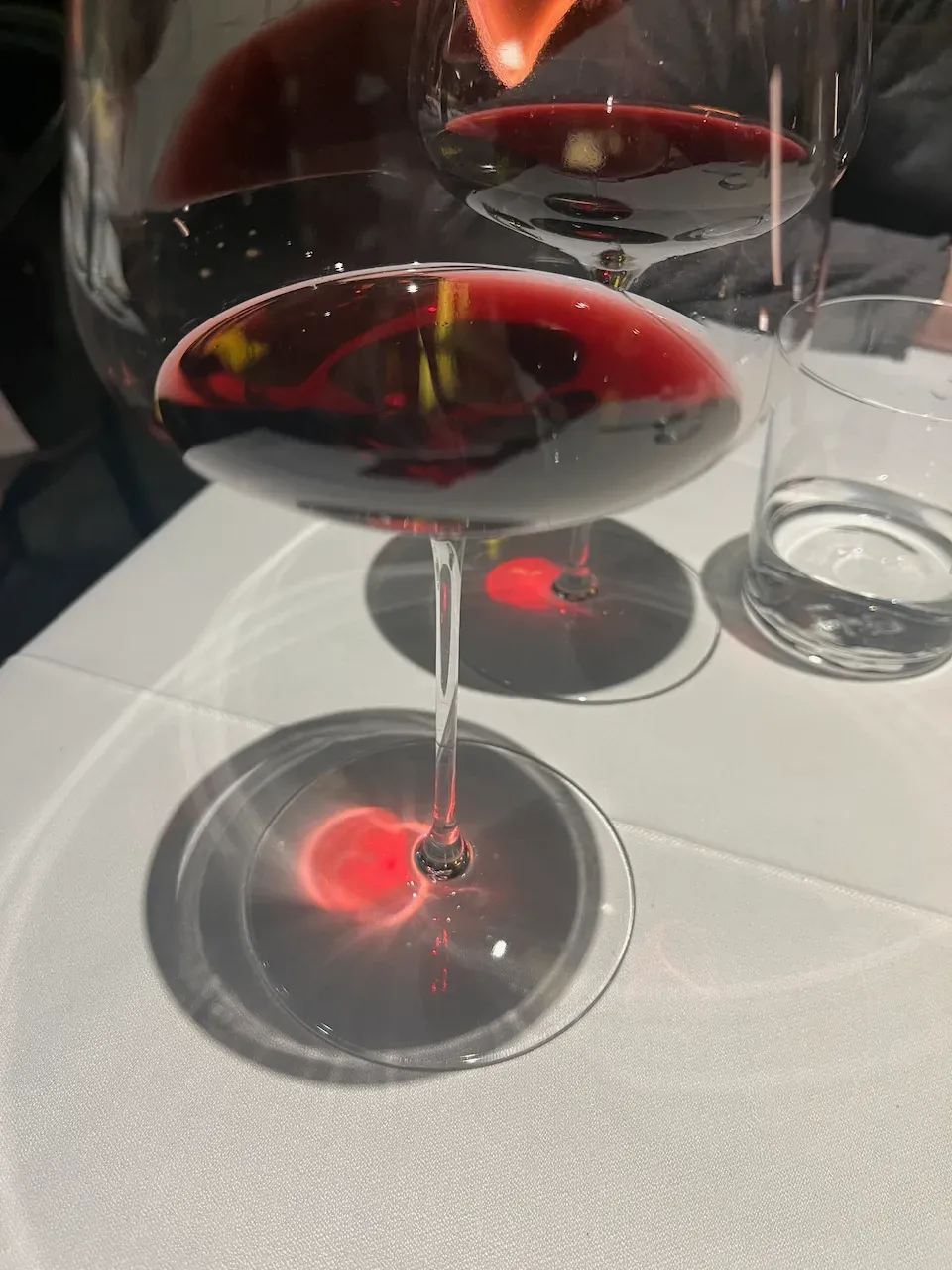 Close-up of a glass of red wine casting a glowing red reflection on a white tablecloth.