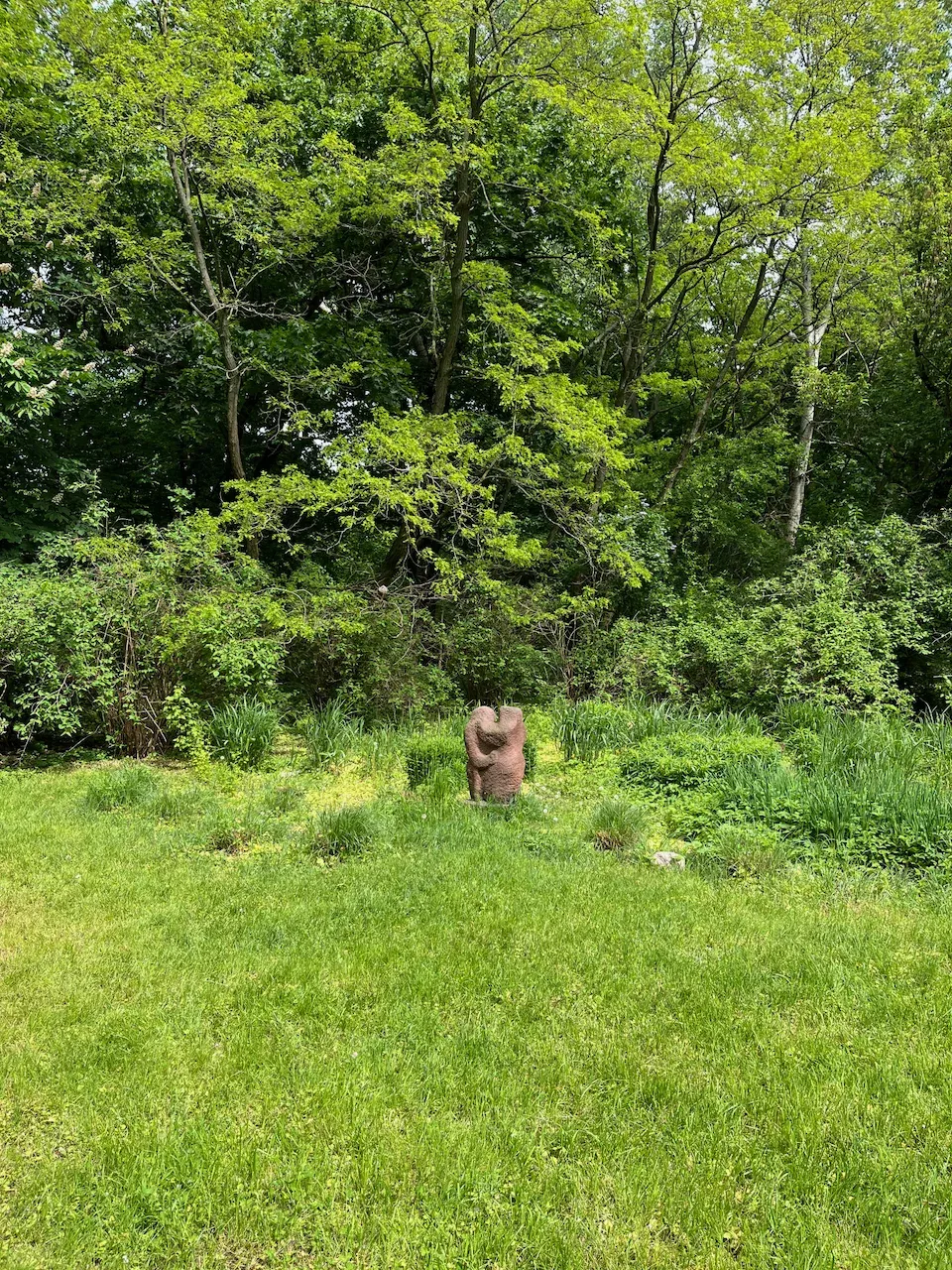 Abstract stone sculpture of two embracing figures in a green forest clearing.