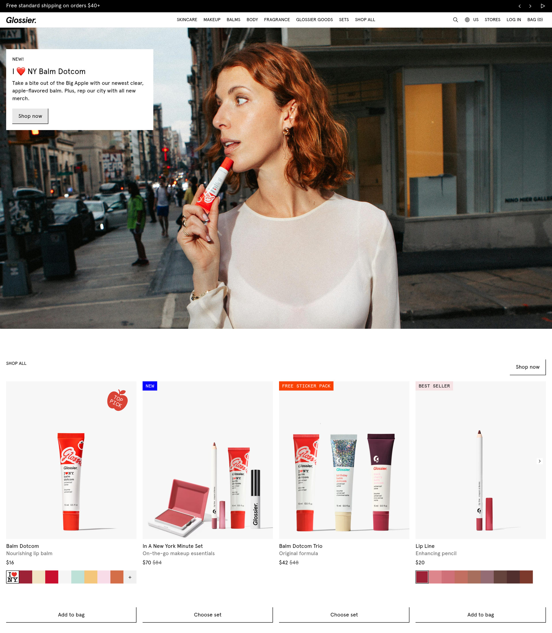 Glossier homepage: a real person applying Balm Dotcom on a New York street, with a shop-all product grid below