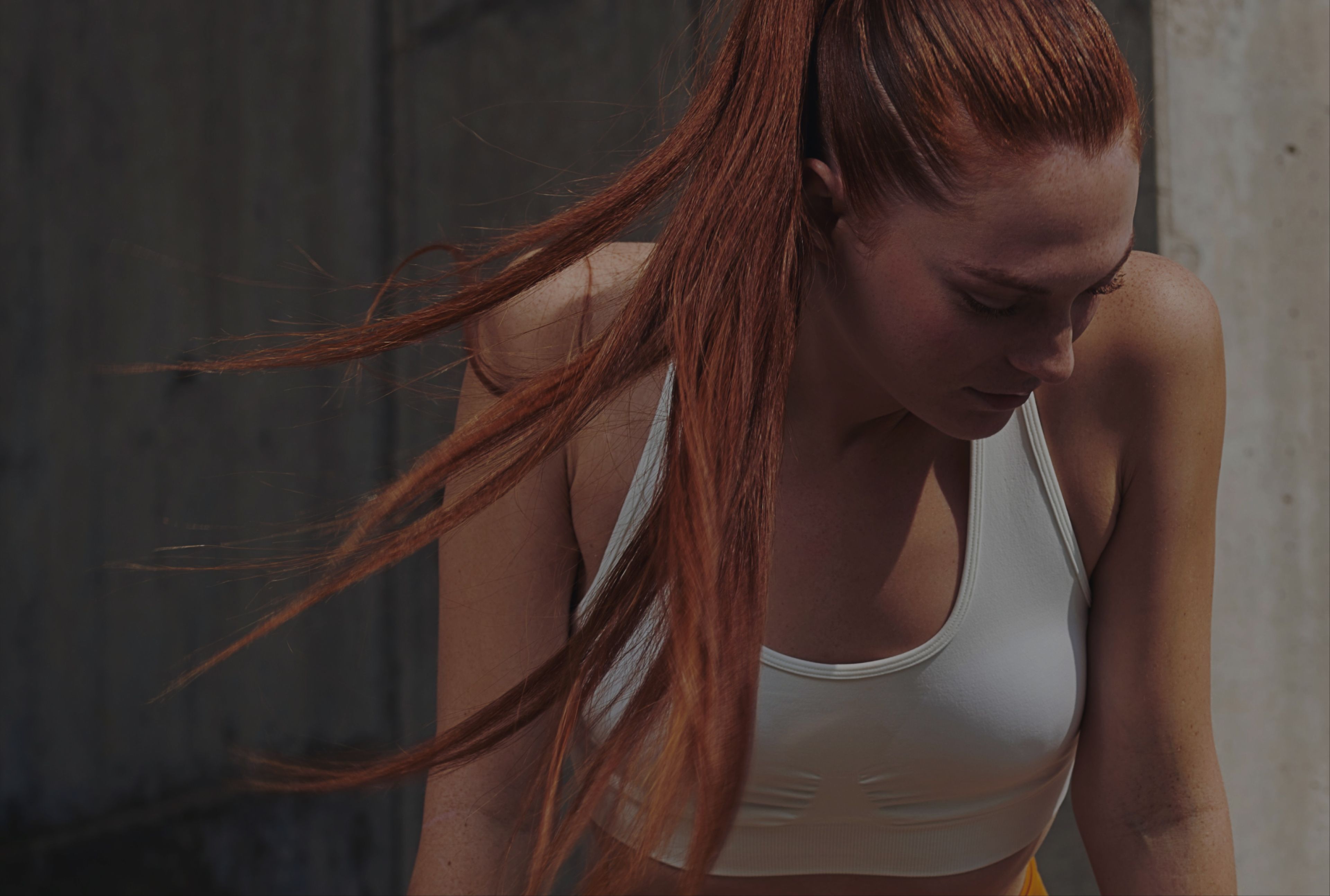 Side profile of a woman with long red hair in a ponytail, wearing a white sports bra and leaning forward in a contemplative pose outdoors.