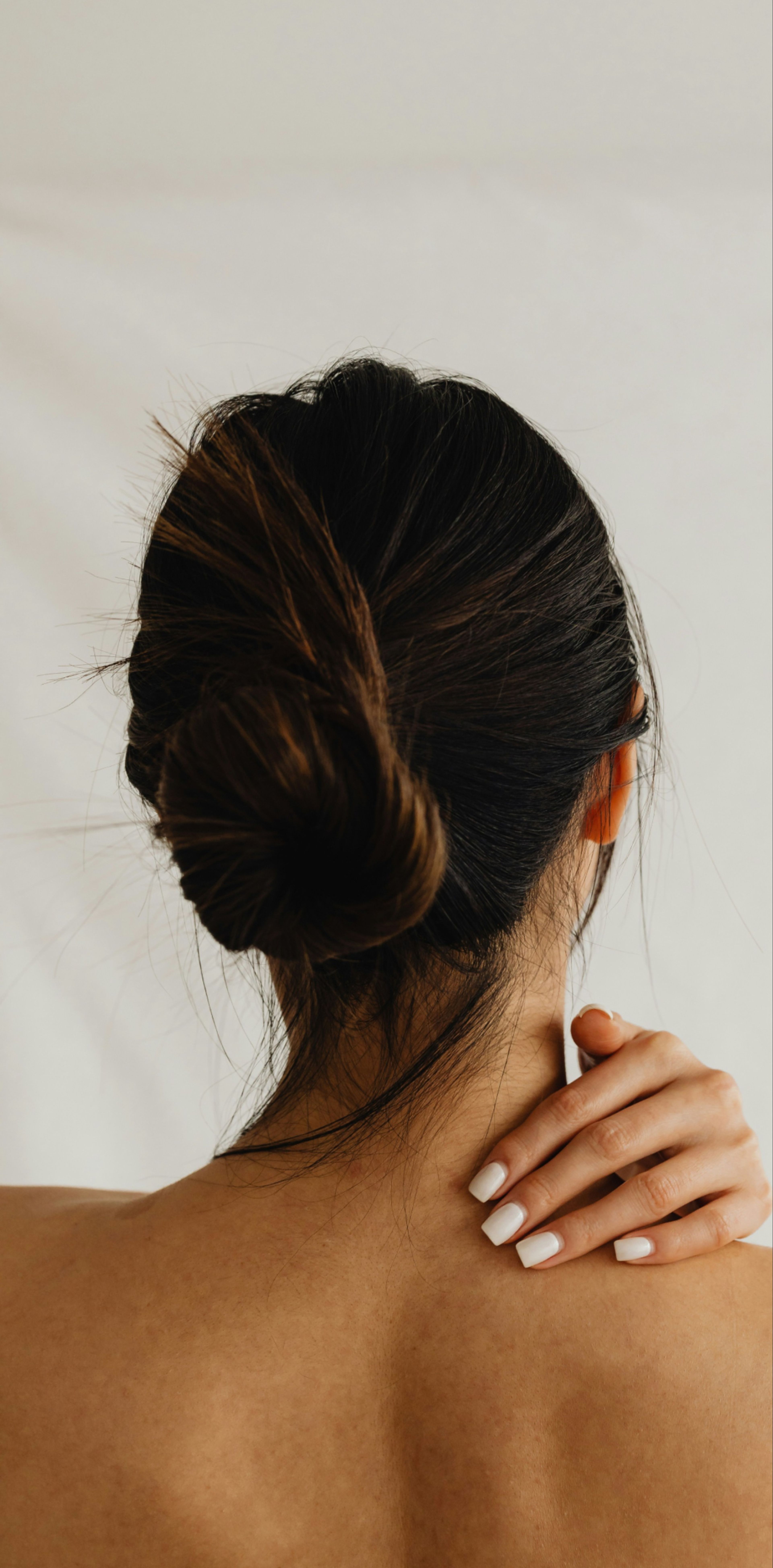 A person with dark hair tied in a bun viewed from behind, gently touching their shoulder with manicured white nails.