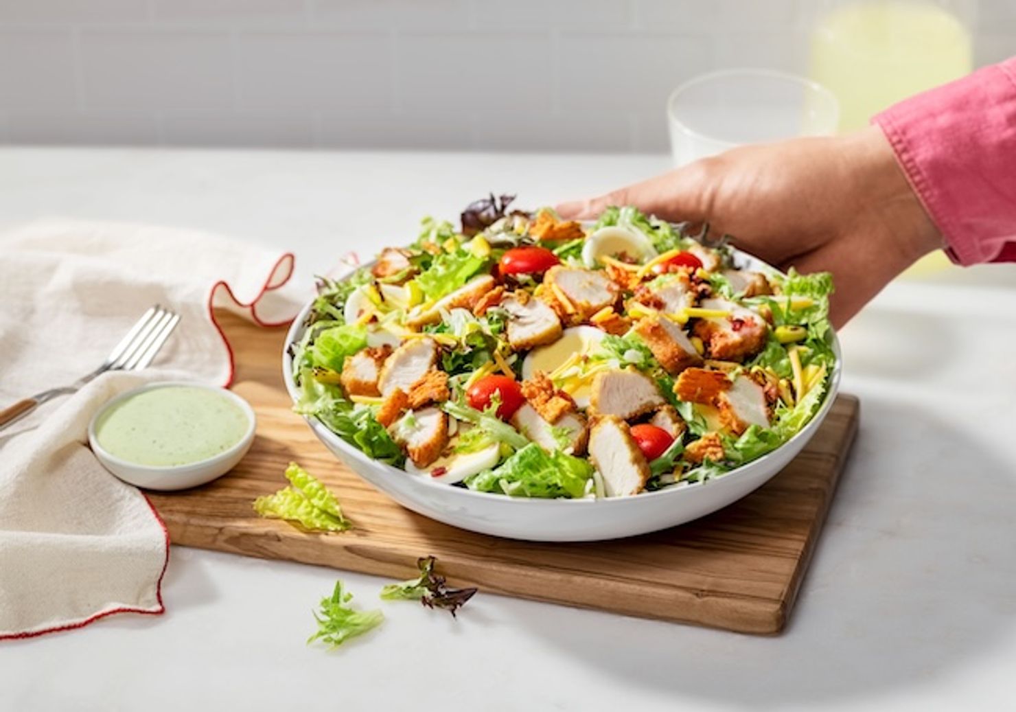 Person placing a Cobb Salad on a cutting board next to a small bowl of Avocado Lime Ranch dressing, a fork and a napkin