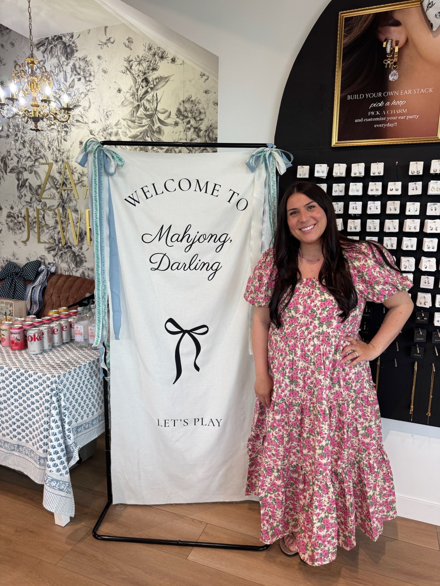 A smiling woman in a floral dress beside the Mahjong Darling welcome sign