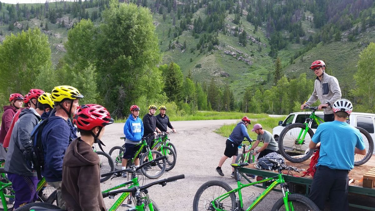 Young riders in helmets on green mountain bikes gathered around a leader, with a wooded hillside behind them