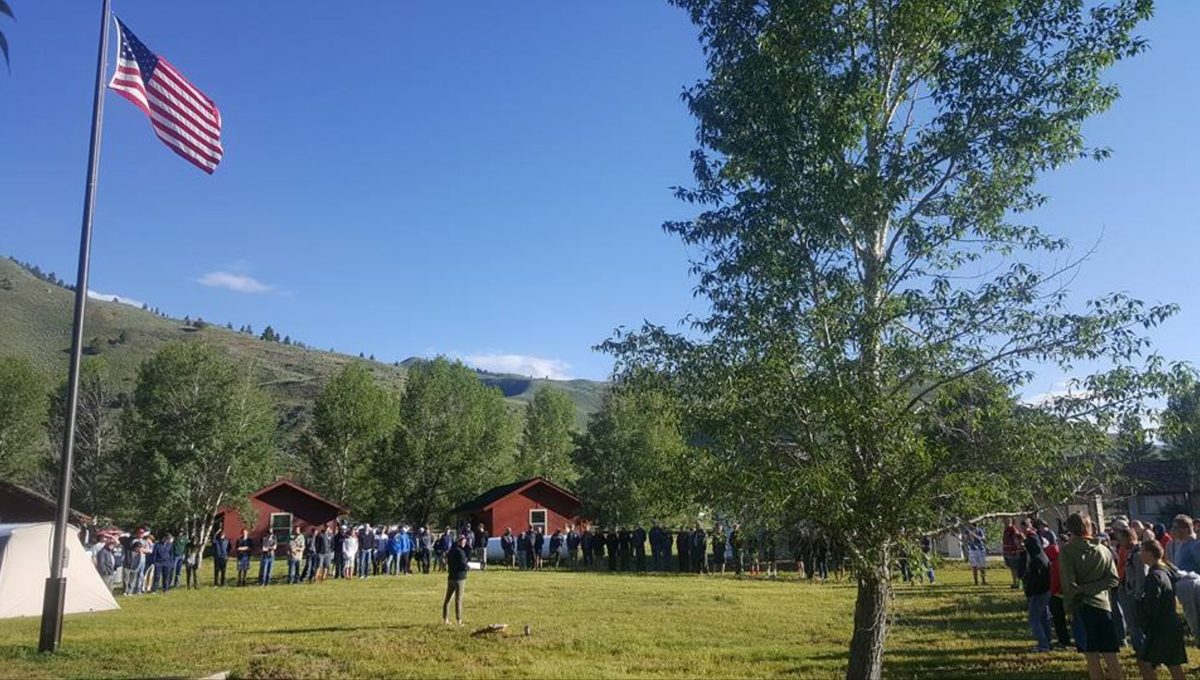 Campers gathered in a circle on the lawn beneath the flag, with red cabins behind them