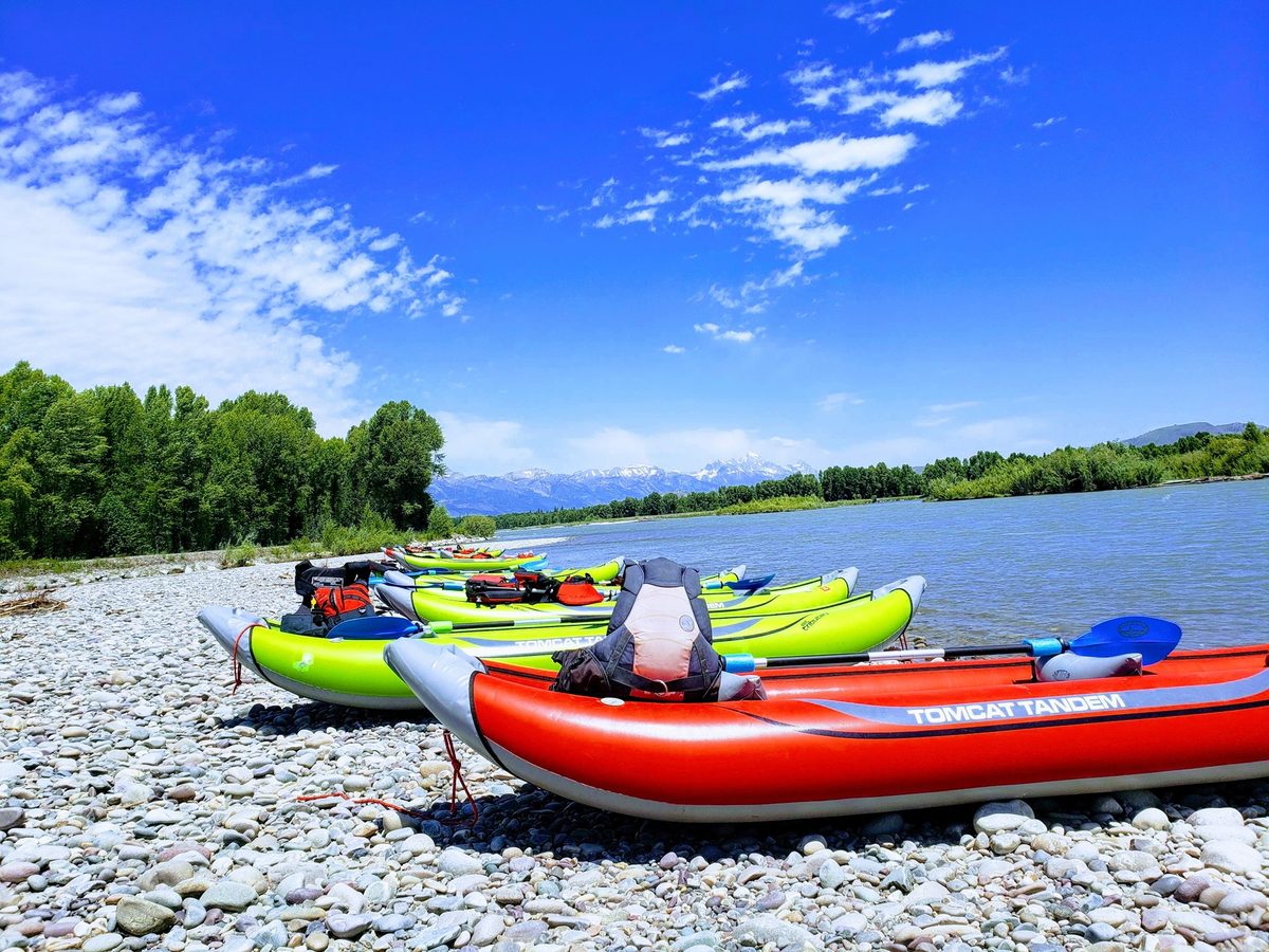Red and green inflatable kayaks lined up on a gravel bar beside the river, with snow-capped mountains in the distance