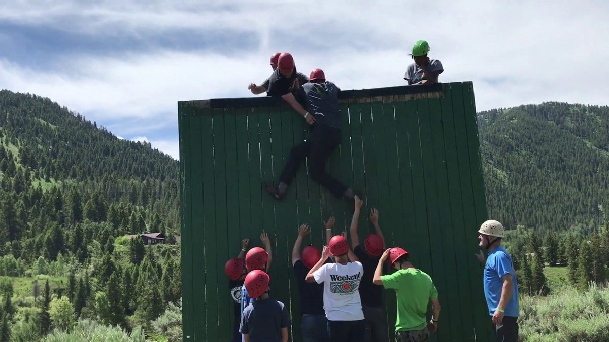 Campers in helmets boosting a teammate over a tall wooden wall