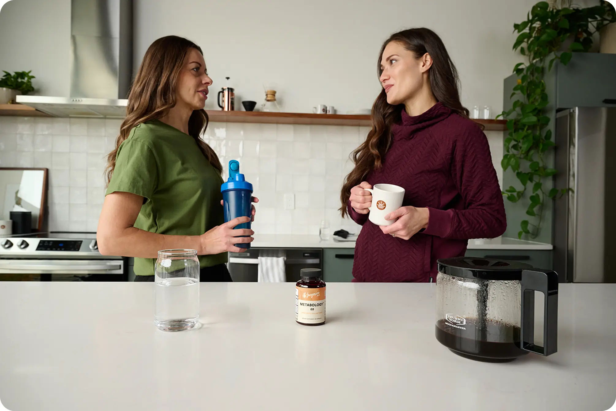 two women talking while holding various health and fitness items in a kitchen.