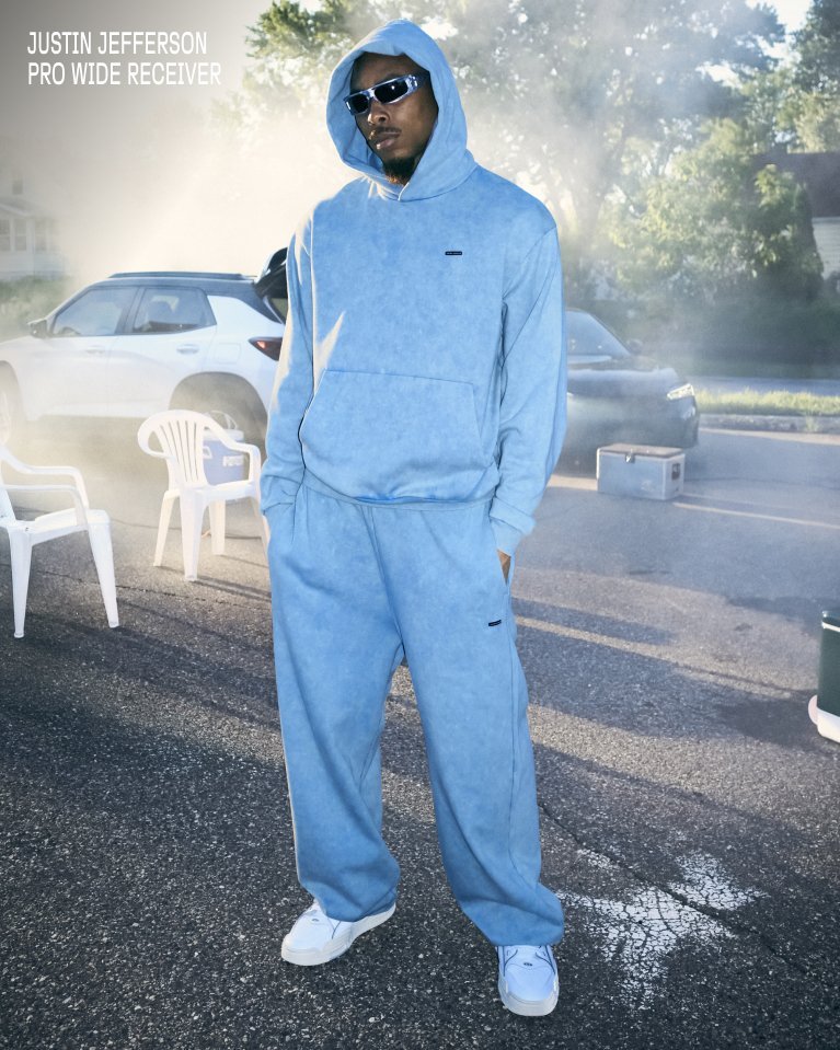 Athlete in light blue Under Armour hoodie and sweatpants stands on smoky street wearing sunglasses.